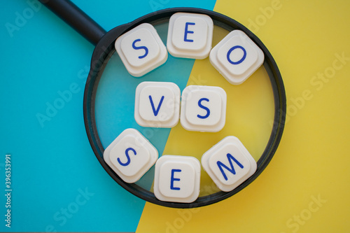 "SEO vs SEM" words over a magnifying glass, on a blue and yellow background. Search engines concepts