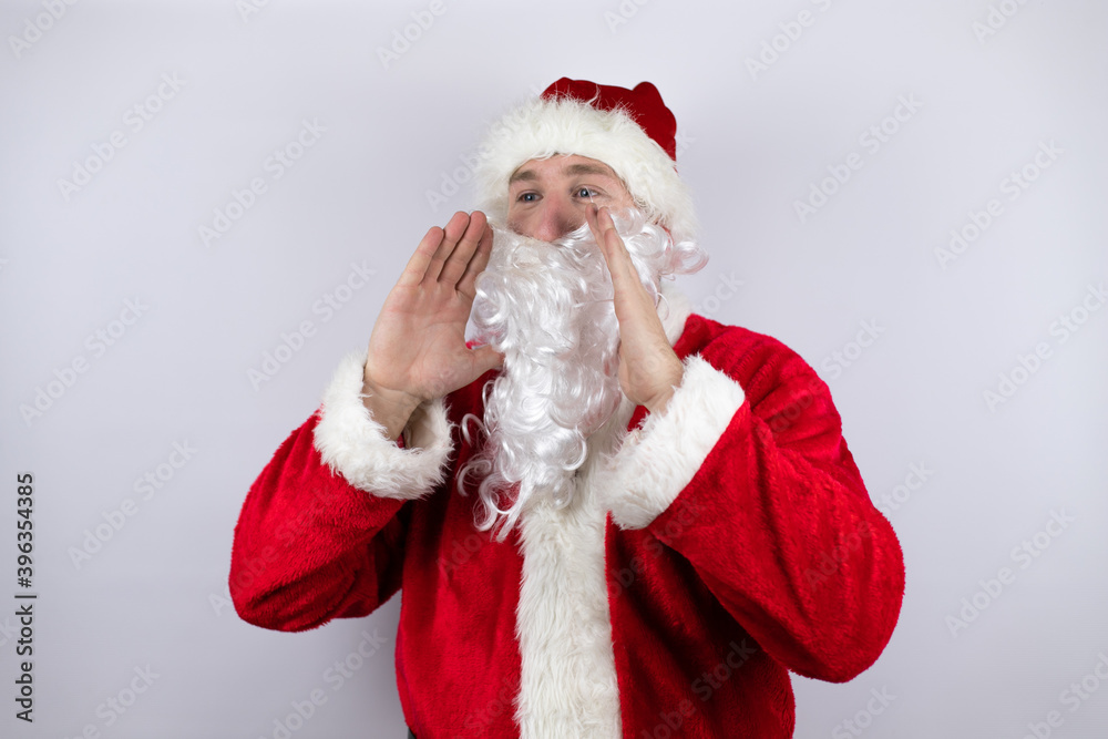 Man dressed as Santa Claus standing over isolated white background shouting and screaming loud to side with hands on mouth Man dressed as Santa Claus standing over isolated white background shouting and screaming loud to side with hands on mouth