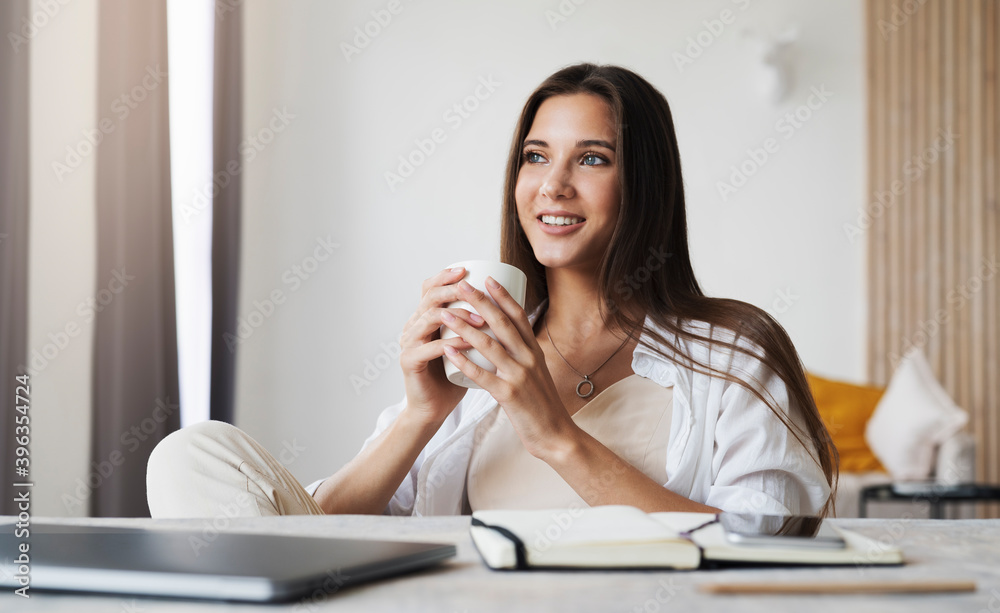 Beautiful brunette girl sits at table in front of laptop, watches television series, holds cup of coffee with hands. Beautiful brunette girl sits at table in front of laptop, watches television series, holds cup of coffee with hands.