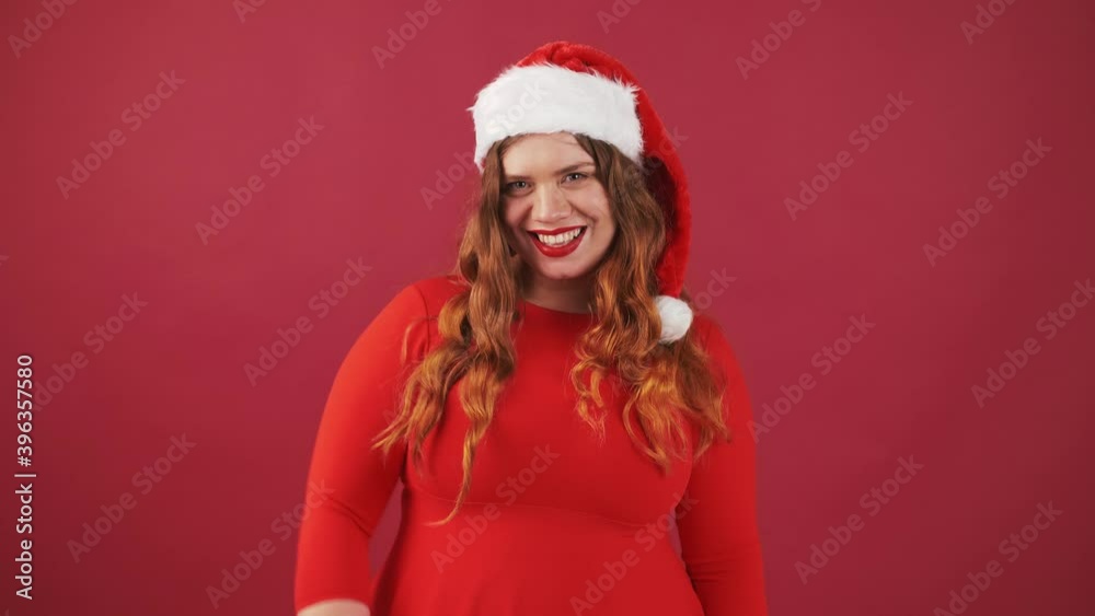 Smiling plus size lady in Christmas red hat, waving hands in hello gesture, greeting friends, red studio background. Smiling plus size lady in Christmas red hat, waving hands in hello gesture, greeting friends, red studio background.