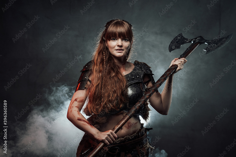 Wielding axe smiley woman viking dressed in dark light armour with brown hairs poses in dark foggy background looking at camera.