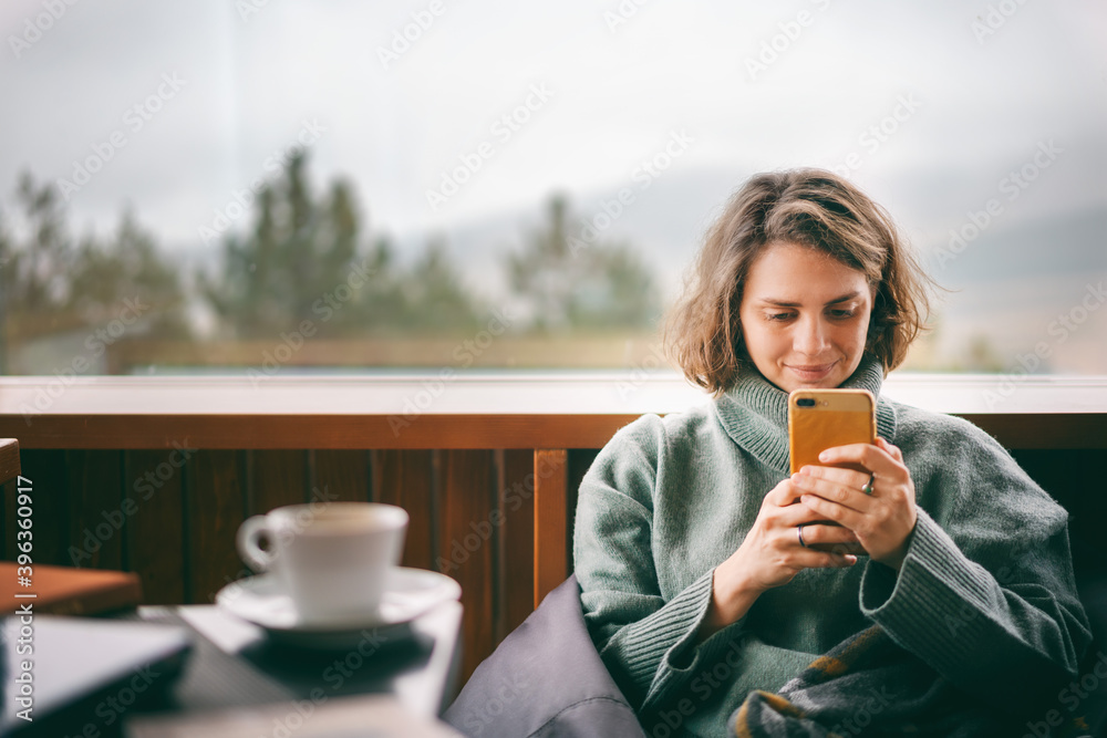 Young woman in a green sweater using a mobile phone near window in country house with a view of the mountains and pine trees