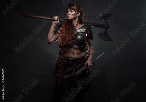 Barbaric and grimy woman dressed in dark armour holding two axes and posing in dark background. © Fxquadro