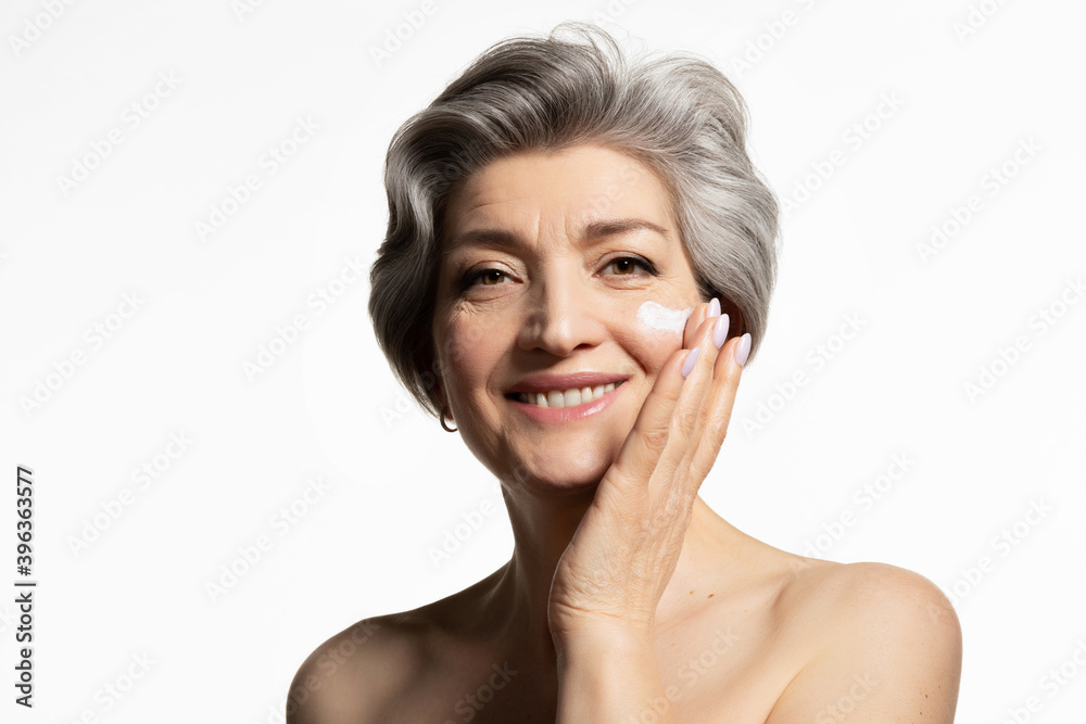 Senior woman putting cosmetic moisturizing rejuvenating creme on face. Smiling elderly 60s female with naked shoulders, close up studio portrait isolated on white. Anti-wrinkle skincare