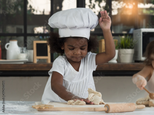 Adorable dark skinned African girl wearing chef hat playing with baking dough in the kitchen at home.