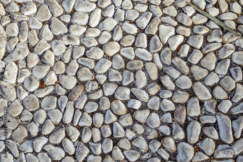 Texture of white sea pebbles cobbled on the sidewalk, close-up. Real pattern of floor paved with natural smooth stones with debris in between, outdoors