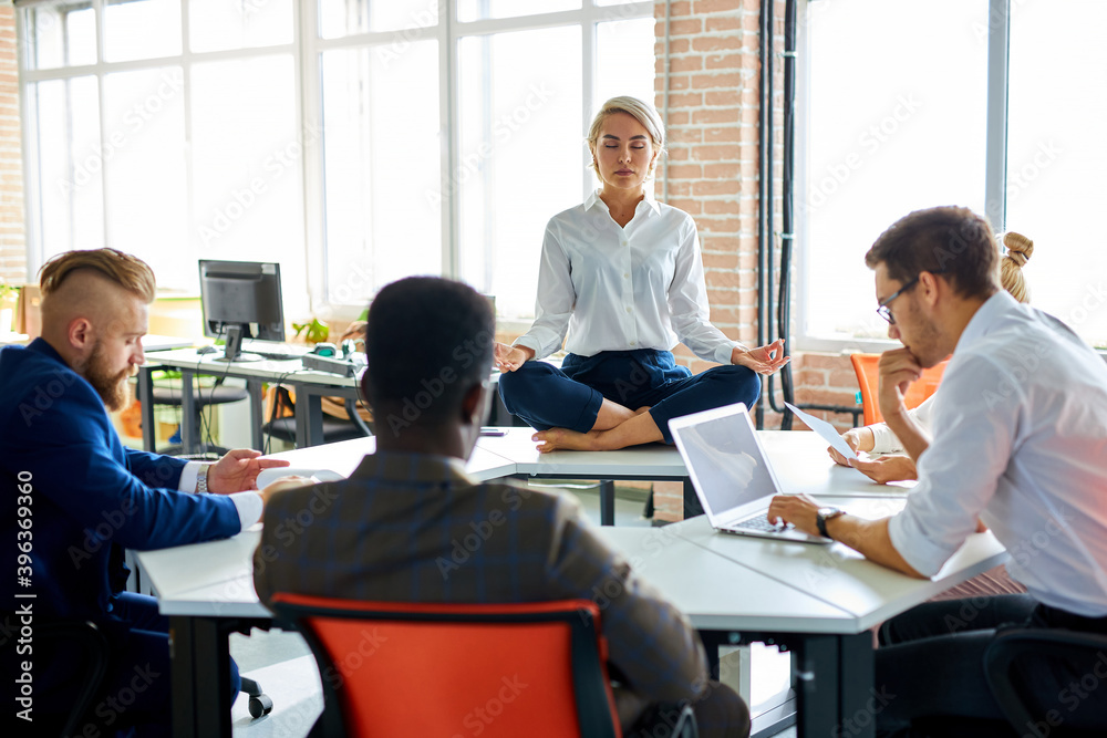 young caucasian woman meditating in office while colleagues are working, keep calm, meditate. sits on table with crossed legs