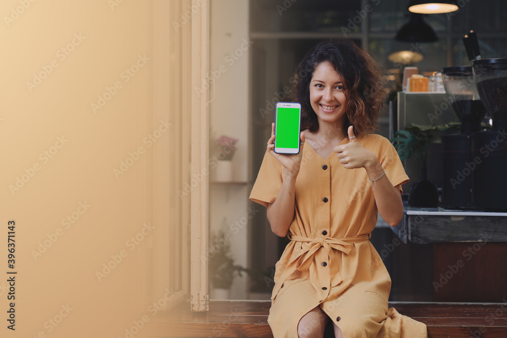 Mockup image of a woman wearing yellow dress holding and showing mobile phone with blank green screen at coffee shop