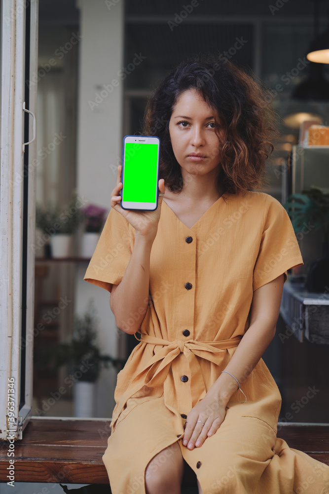 Mockup image of a woman wearing yellow dress holding and showing mobile phone with blank green screen at coffee shop