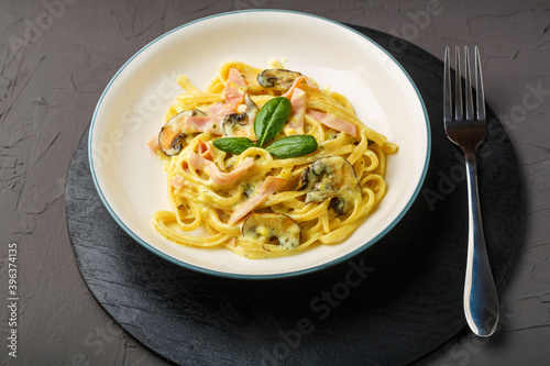 a plate of pasta with ham and mushrooms in a creamy basil sauce on a black round stand with a fork.