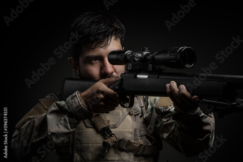 Latin man with a beard and black hair standing, holding a rifle with his two hands, uses an earphone and camouflage clothing, studio with dark background