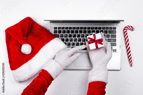 Santa Claus hands in medical latex gloves showing Christmas gift box on laptop with Santa's hat and Christmas candy can on white background.