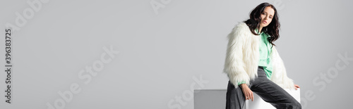  young woman in faux fur jacket posing on cube on white background, banner