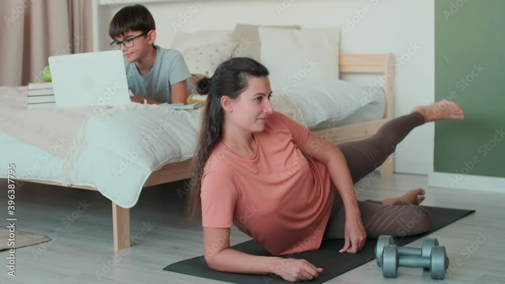 Home Isolation, Distance Learning, Remote Work, Fitness at Home, Online Training, Family Quarantine. Mom goes in for sports at home on the floor next to her son who does homework on the bed Home Isolation, Distance Learning, Remote Work, Fitness at Home, Online Training, Family Quarantine. Mom goes in for sports at home on the floor next to her son who does homework on the bed