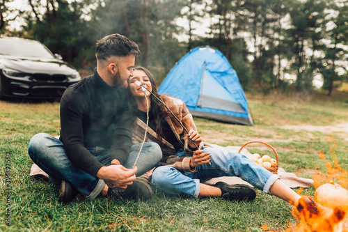 Beautiful couple of lovers with roasting marshmallows in hands spend the weekend in nature with tent near the campfire