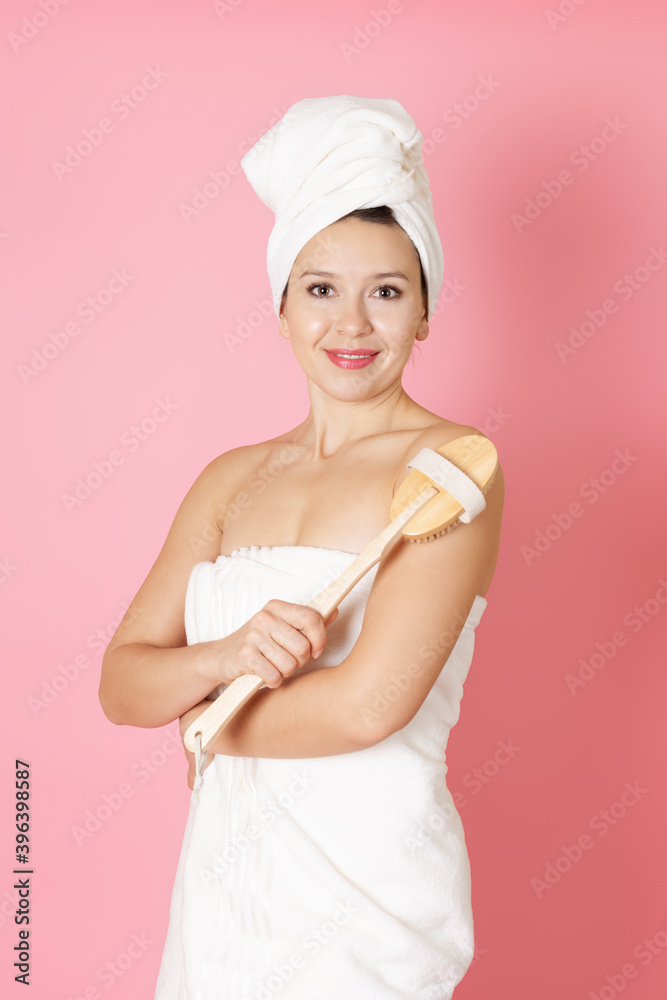 close up portrait of a smiling woman in a towel massaging her body with a wooden brush isolated on a pink background.