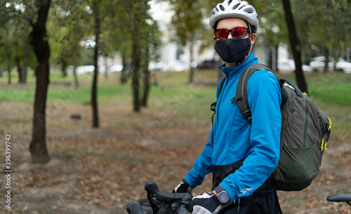 A man of Caucasian nationality in the park with a bicycle in a helmet and a protective mask. Food delivery and outdoor sports in quarantine.