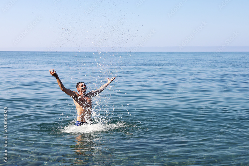 Joyful man splashes in the water, alone at the sea, where there are few tourists resting around