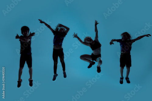 Silhouetted full length shot of four little sportive kids looking joyful while posing, jumping isolated over blue background
