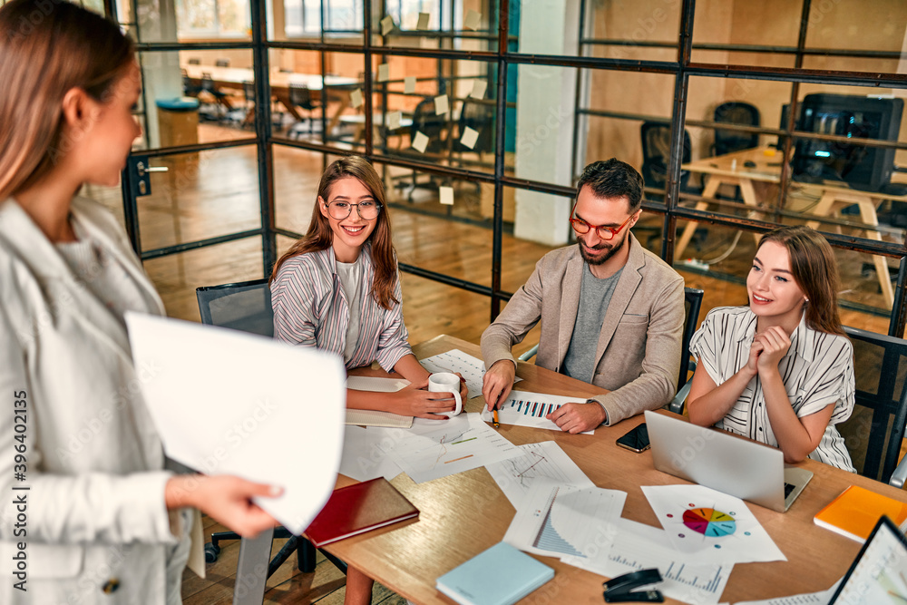 Focused young Caucasian business woman showing graph on paper to group of colleagues sitting at table with laptops in modern office.