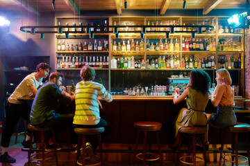 Rear view of three guys drinking beer, looking at women, two girlfriends sitting at the bar counter. Friends spending time at night club, restaurant