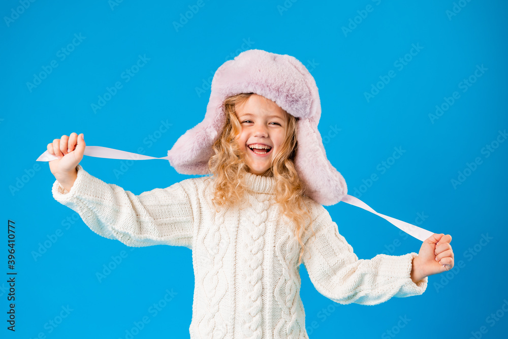 portrait of a little blonde girl in a winter hat and knitted sweater on a blue background. baby girl in winter clothes smiling on blue background in Studio, space for text portrait of a little blonde girl in a winter hat and knitted sweater on a blue background. baby girl in winter clothes smiling on blue background in Studio, space for text
