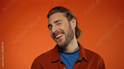 joyful and bearded man winking eye while looking at camera on orange