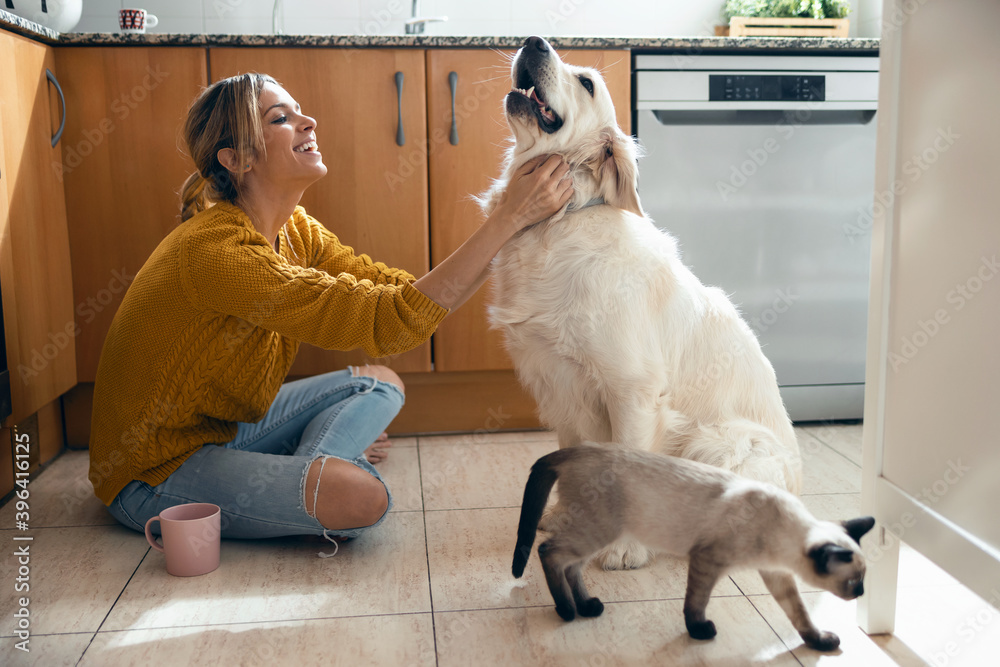 Beautiful young woman stroking her cute lovely dog sitting on the floor in the kitchen at home.