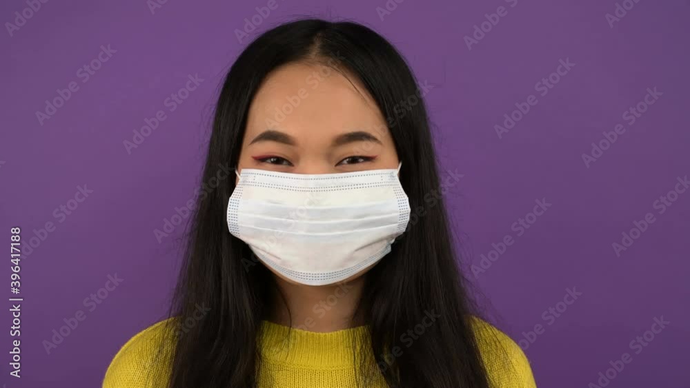 Smiling young Asian woman takes off her protective medical mask. lady in yellow sweater on purple isolated background. 4K Smiling young Asian woman takes off her protective medical mask. lady in yellow sweater on purple isolated background. 4K