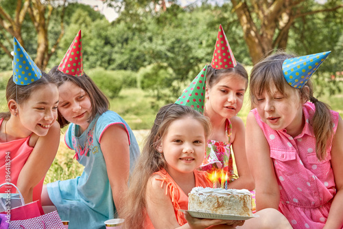 A girl holds a cake with burning candles and looks at the camera. There are many friends nearby. Birthday in nature. Picnic in nature, in the Park in summer.