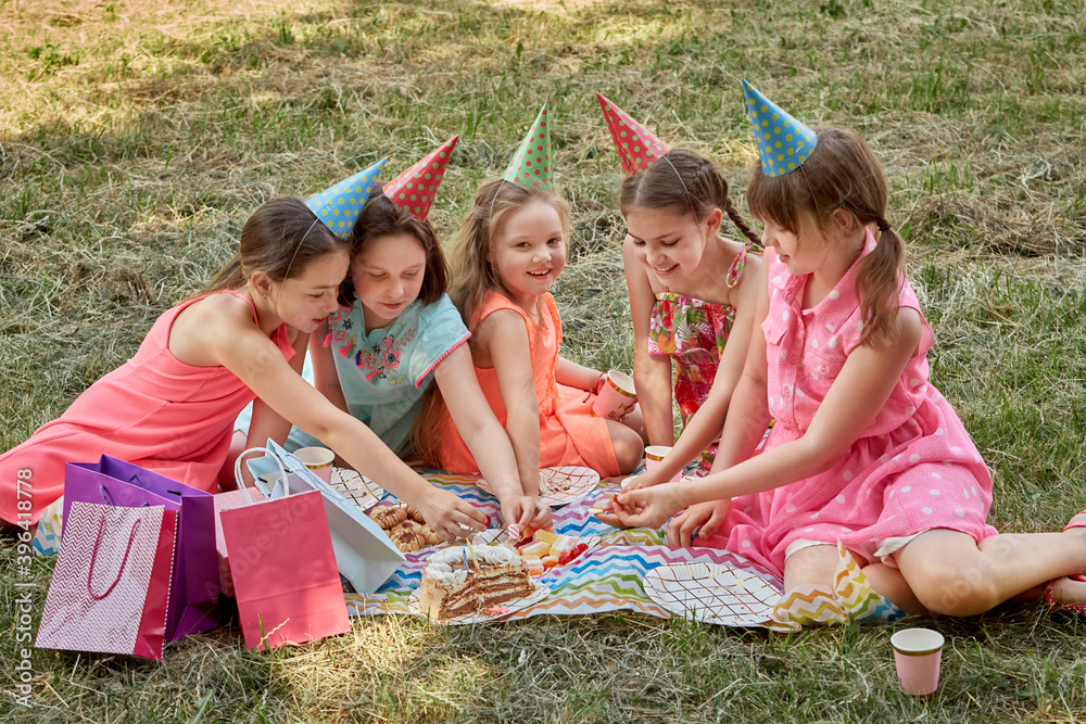 Fototapeta premium Little girls celebrate their birthday in the summer on a picnic. They eat sweets. A lot of gifts in festive bright packages.