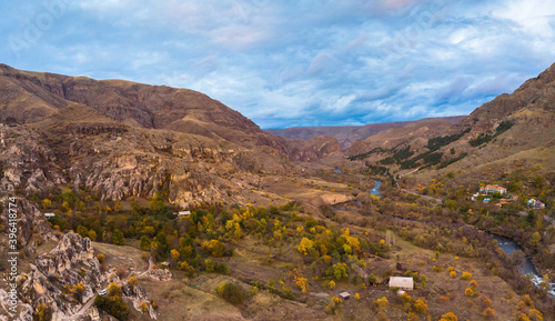 HDR panorama view of Vardzia canyon surounded by autumn landscape and rocky cliffs and few houses in foreground.