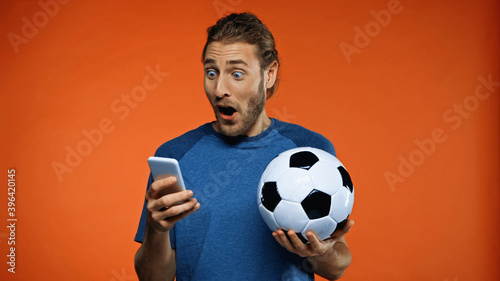 shocked football fan in blue t-shirt using smartphone and holding soccer ball on orange © LIGHTFIELD STUDIOS
