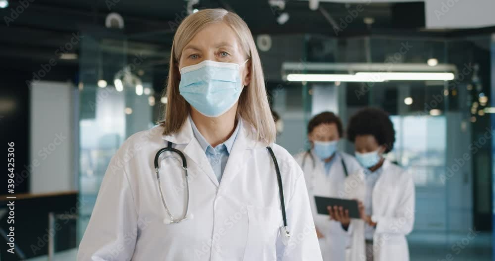 Close up portrait of Caucasian senior female doctor in medical mask standing in hospital looking at camera at work. Healthcare professionals using tablet on background. Coronavirus concept Close up portrait of Caucasian senior female doctor in medical mask standing in hospital looking at camera at work. Healthcare professionals using tablet on background. Coronavirus concept