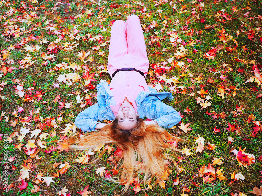 A red-haired girl in a pink overalls lies on a meadow covered with autumn maple leaves