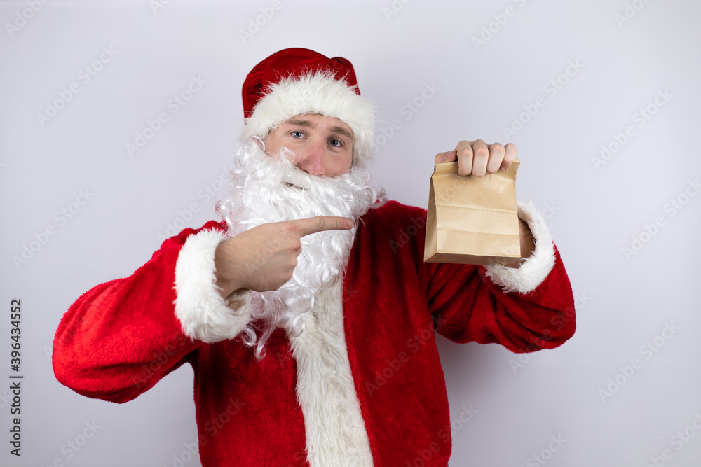 Man dressed as Santa Claus standing over isolated white background holding a paper bag and pointing the bag Man dressed as Santa Claus standing over isolated white background holding a paper bag and pointing the bag