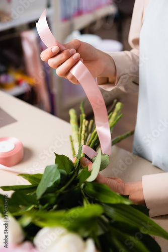 Close up view of florist hand holding decorative ribbon, while tying stalks of bouquet in flower shop on blurred background