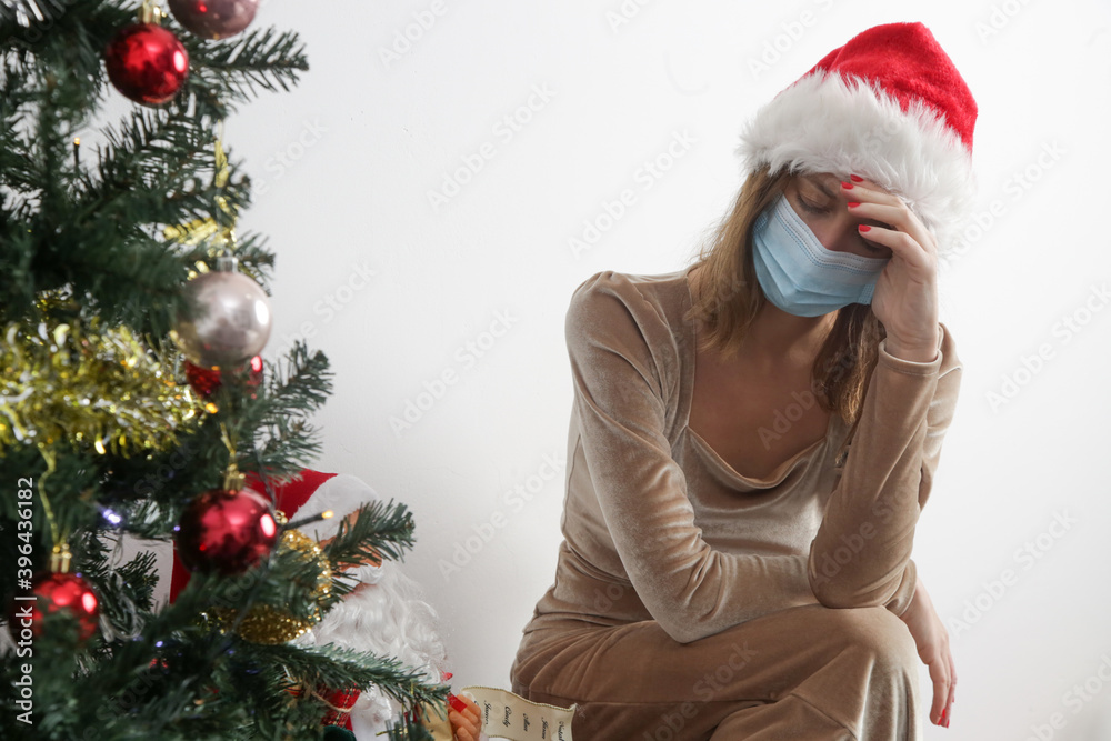 Young stressed woman wearing proetctive medical mask and Santa hat next to the Christmas tree at home. Winter holidays during Corona virus outbreak, self-quarantine concept. Young stressed woman wearing proetctive medical mask and Santa hat next to the Christmas tree at home. Winter holidays during Corona virus outbreak, self-quarantine concept.