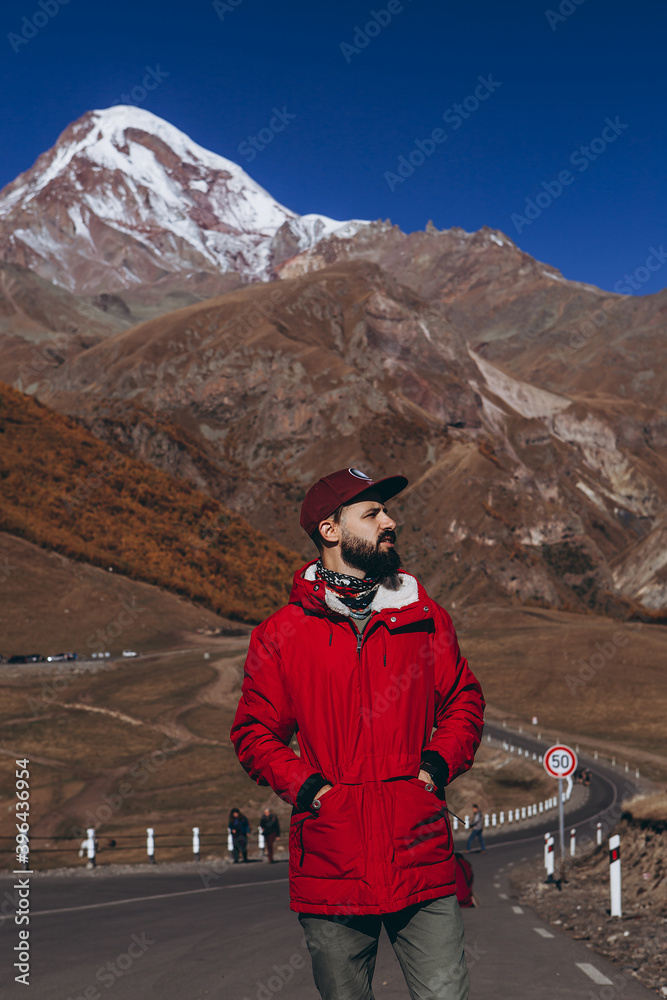 young attractive man with a beard dressed in a red jacket climbs the asphalt road on the slopes of the Caucasus Mountains against the backdrop of Mount Kazbek young attractive man with a beard dressed in a red jacket climbs the asphalt road on the slopes of the Caucasus Mountains against the backdrop of Mount Kazbek
