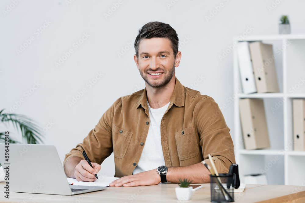 Fototapeta premium Smiling businessman looking at camera while writing in notebook at workplace with laptop on blurred background