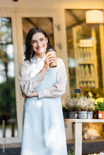 smiling florist holding coffee to go near flower shop and potted plants on blurred background