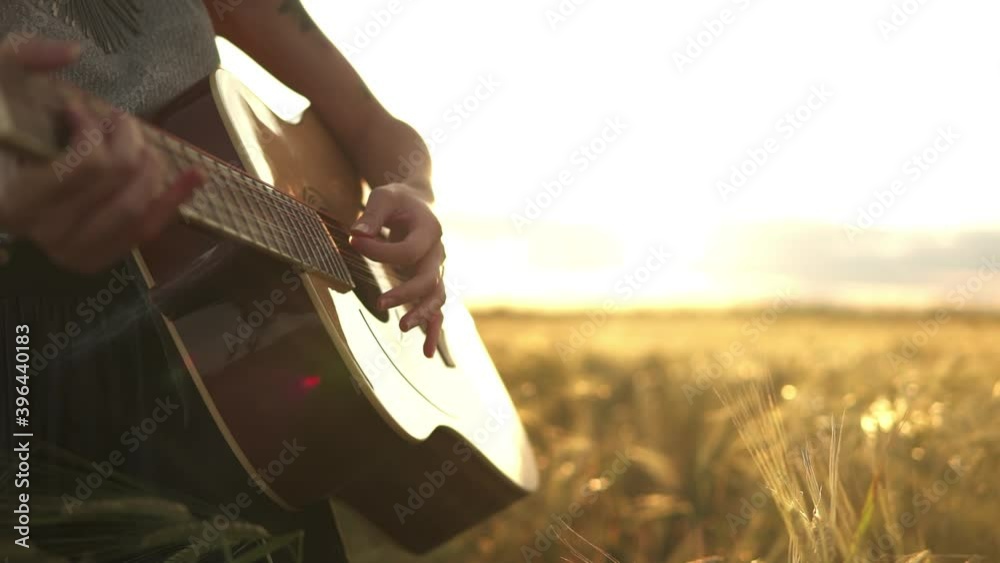 Young female person playing music on acoustic guitar and singing outdoors in summer. Musician guitarist lady play in meadow field at sunset. Close-up hands. People, lifestyle, expression concept.