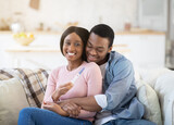 Young African American family learning about their future child from pregnancy test at home, embracing on sofa