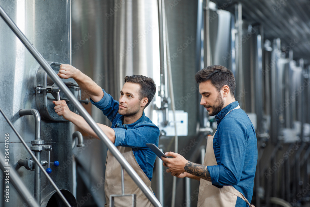 Millennial guy worker in apron turns a valve on big boiler for lager, manager with tablet controls process in interior