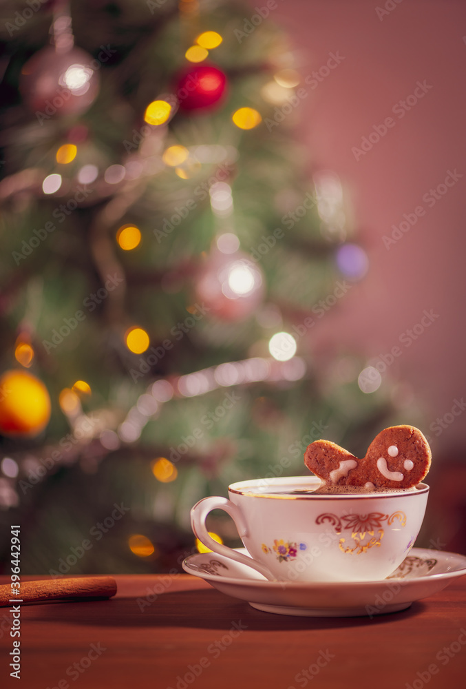 Fototapeta premium Gingerbread man cookie in a coffee mug in front of decorated Christmas tree. Christmas scene with sweet gingerbread cookie.