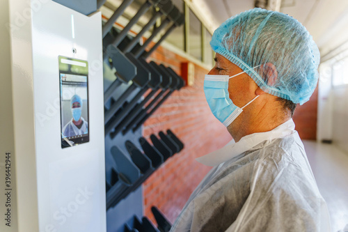 Side view of caucasian man doctor scientist wearing protective gear standing in front of electronic face recognition terminal with digital temperature control camera checking in before going to work photo