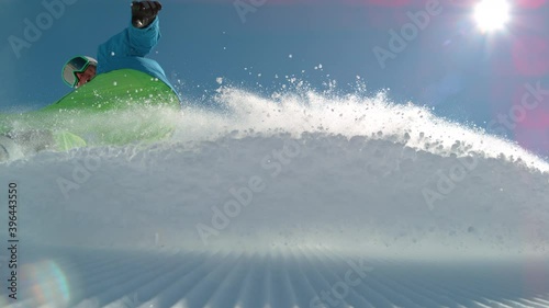 SLOW MOTION TIME WARP, LOW ANGLE, CLOSE UP, DOF: Athletic male tourist shredding the slopes sprays snow at the camera. Snowboarder carves along a groomed ski slope of a ski resort in the sunny Alps.
