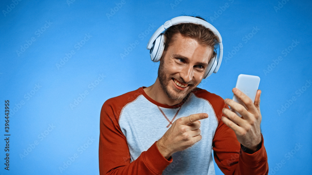 happy man in wireless headphones pointing with finger at phone on blue happy man in wireless headphones pointing with finger at phone on blue