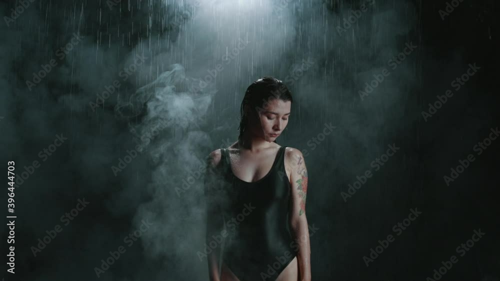 Woman standing alone under rain in smoke isolated on black background and raising hands up. Mind is completely calm. Swimmer overcame the fear of failure, loneliness and resentment, no more tears Woman standing alone under rain in smoke isolated on black background and raising hands up. Mind is completely calm. Swimmer overcame the fear of failure, loneliness and resentment, no more tears