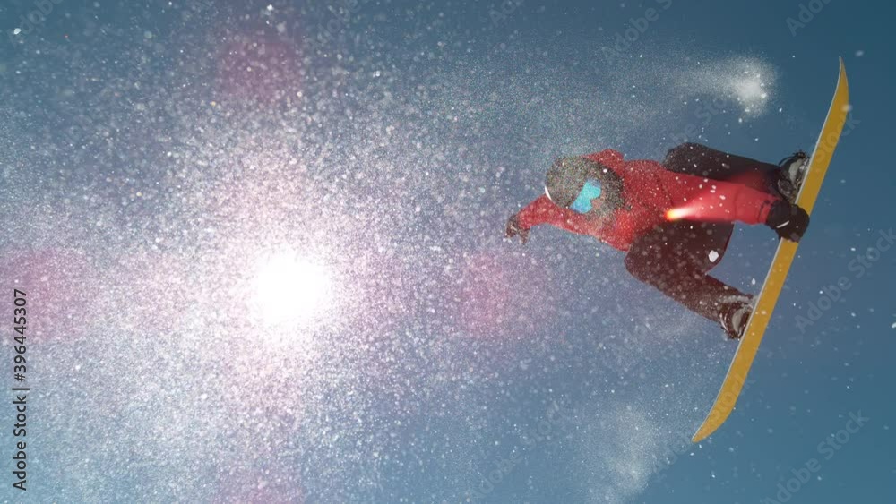 SLOW MOTION TIME WARP, LENS FLARE, BOTTOM UP: Snowflakes sparkle in the sunshine as male rider does snowboarding tricks. Fearless snowboarder does a spin trick while riding off a big air kicker.